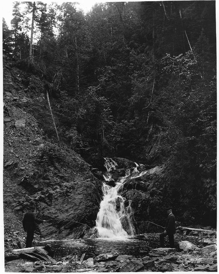 Black and white photograph of two men in suits fishing for trout at the foot of a waterfall. Both banks of the stream are rocky. In the background a steep rocky cliff is partially covered by a forest.