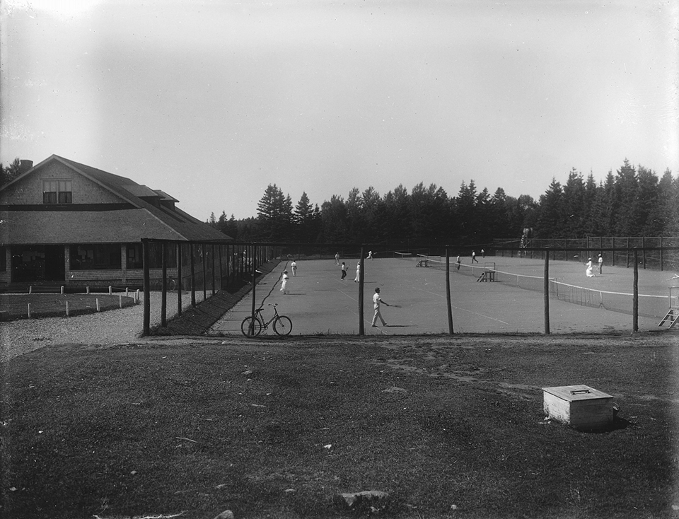 Black and white photograph of the tennis courts at the Cascade Golf and Tennis Club. A high wooden fence surrounds the tennis court. Men, wearing white trousers and a white shirt, and women, wearing a long white skirt in a white shirt with long sleeves and wearing a hat, play tennis. On the left side is the club house.