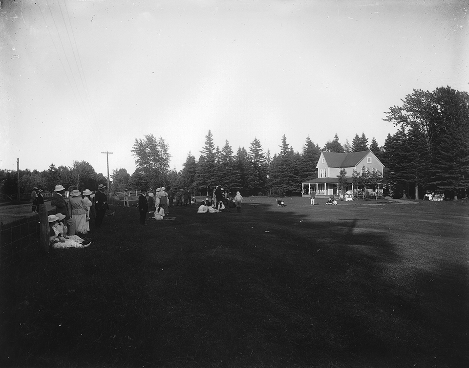 Black and white photograph of the first tee of the Cascade Golf Club. Twenty individuals dressed in the fashion of 1914 watch and wait. Some women are sitting on the grass. In the background, a two-storey cottage with clean Victorian architecture.