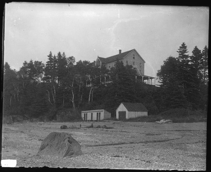 Black and white photograph of the Cascade hotel. The three-storey wooden hotel is located at the top of a steep cliff. The cliff is covered with a forest of spruce and white birch trees. At the foot of the cliff is the rocky beach of Metis. Between the bottom of the cliff and the beginning of the beach there is a small boathouse and changing cabins. Both buildings are made of wood.