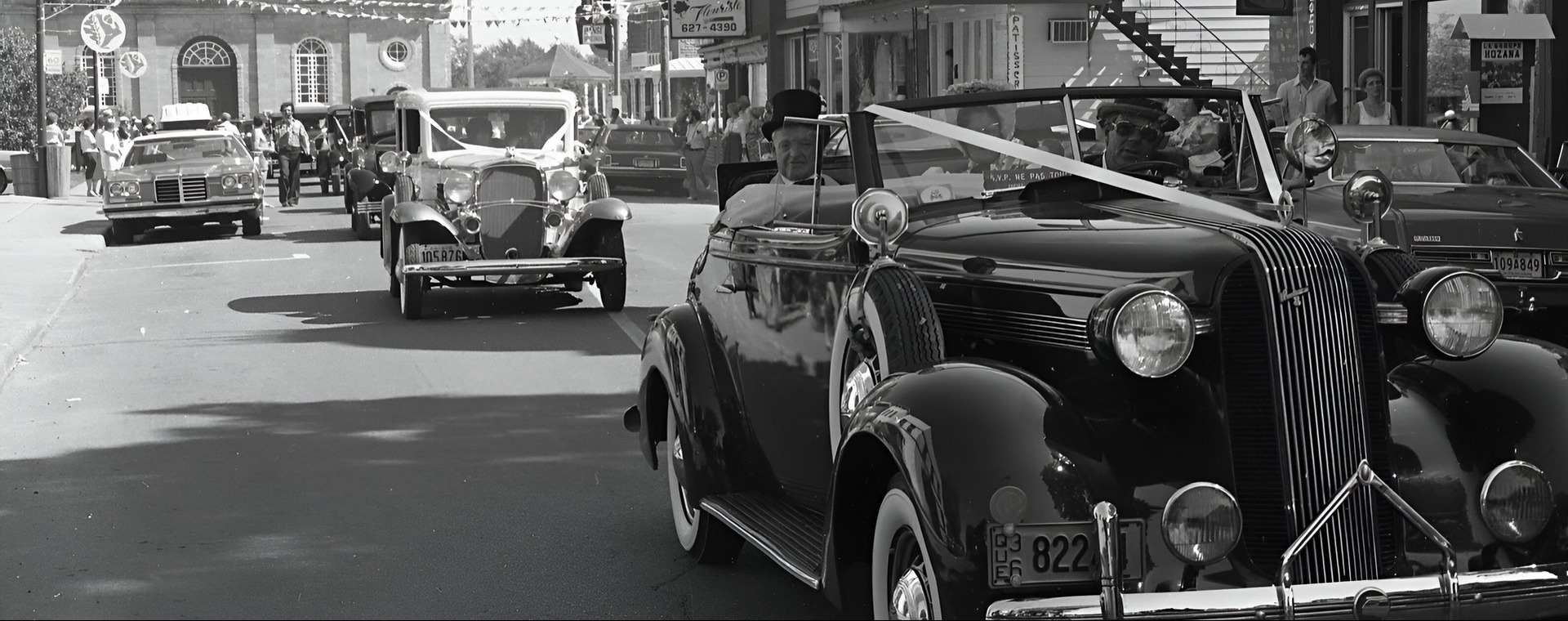 A black and white photograph of cars driving down Rue Saint-Eustache. Shops can be seen on the right side of the street, and the Saint-Eustache church is in the background.