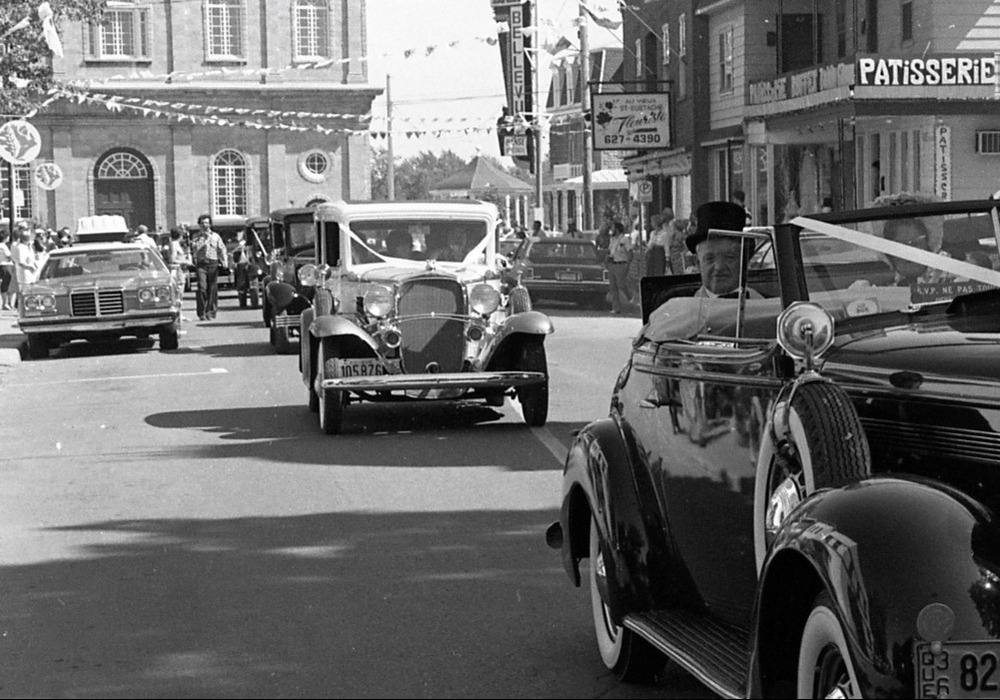 A black and white photograph of cars driving down Rue Saint-Eustache. Shops can be seen on the right side of the street, and the Saint-Eustache church is in the background.