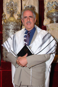A man wearing a suit and white prayer shawl, standing in front of Torah scrolls in a synagogue ark.