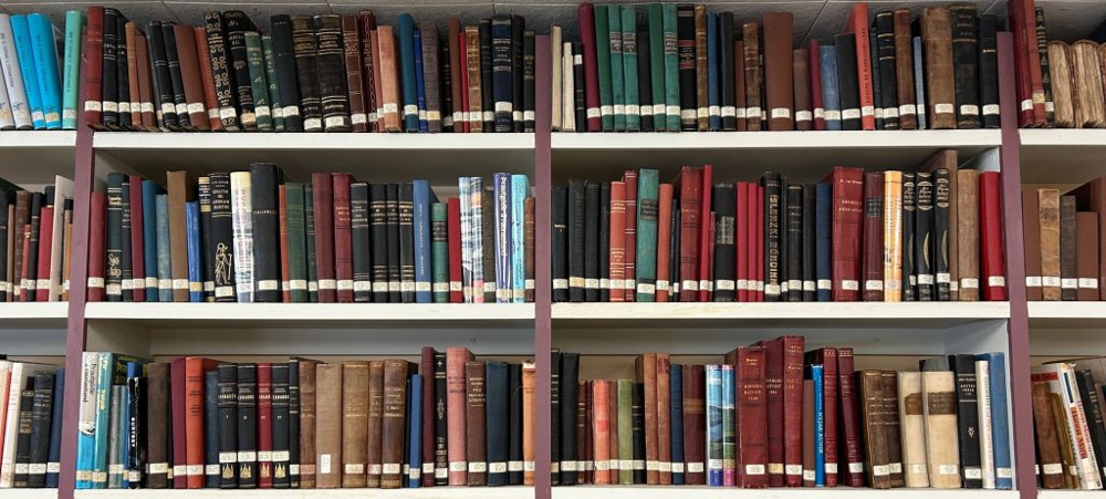 A photo of three tall shelves filled with tightly packed books in a library or archive. Most of the books are older and bound in a variety of rich colors such as red, green, brown, and black, with white spine labels indicating cataloging information. Some titles are in Icelandic. The books appear well-preserved.
