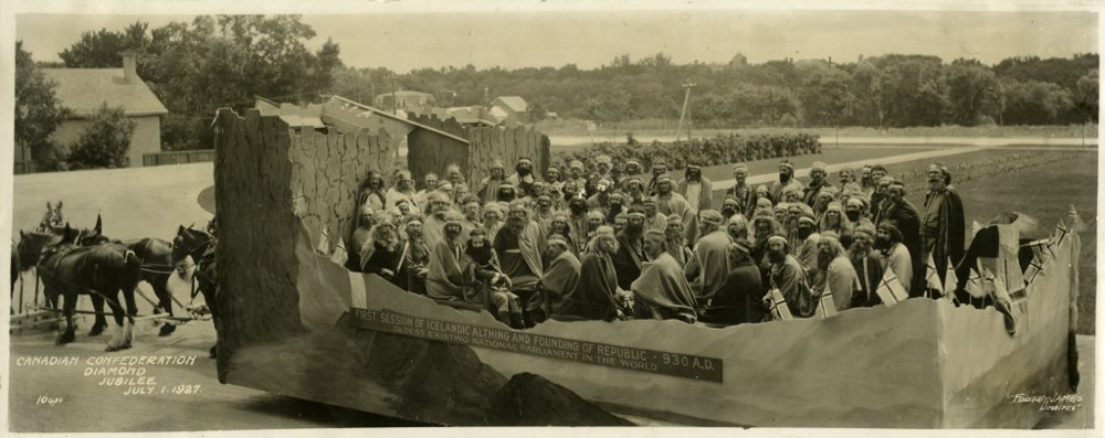 A black-and-white photo of a large parade float from July 1, 1927, featuring dozens of people dressed as early Icelandic settlers. The float is shaped like a large Viking meeting hall and drawn by horses. A banner on the side reads: First Session of Icelandic Althing and Founding of Republic, 930 A.D. – Oldest Existing National Parliament in the World. The image is labeled Canadian Confederation Diamond Jubilee, July 1, 1927.