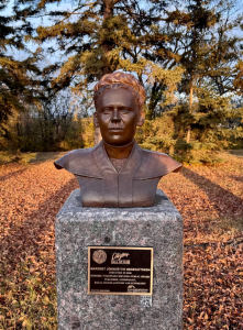 Bronze bust of a woman mounted on a polished stone pedestal set in a park with autumn leaves covering the ground. Behind the statue are tall evergreen and bare trees lit by the warm light of the setting sun. A plaque on the pedestal displays text and logos, indicating this is a commemorative monument. The bust features short hair and a composed expression, facing directly forward.