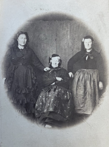A sepia-toned historical portrait showing three Icelandic women. Torfhildur Þorsteinsdóttir Holm stands on the left in a dark, ruffled dress; her mother Guðríður Torfadóttir sits in the center, wearing a patterned skirt and dark jacket; and her sister Ragnhildur Þorsteinsdóttir stands on the right in a simple blouse and apron. The photo has an oval vignette effect and reflects 19th-century Icelandic attire.