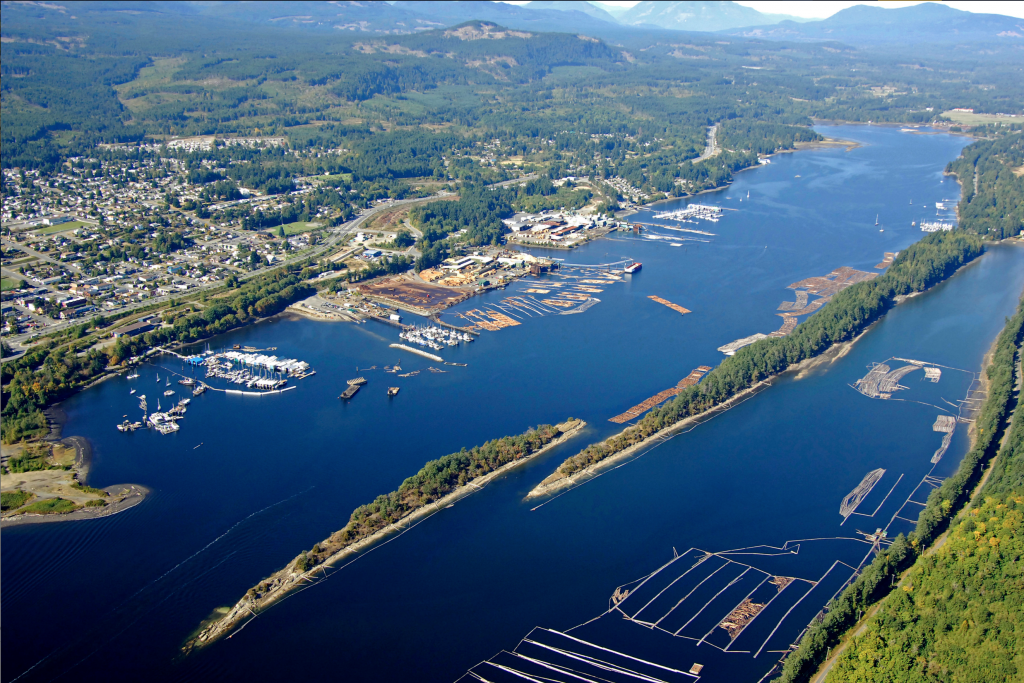 Colour oblique aerial photograph of a town on the shore of an elongate narrow body of water with wooded islands which is bordered by forested hills.