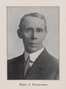 A black and white photo of a middle aged man taken from the shoulders up. He is wearing glasses and a suit jacket and tie.