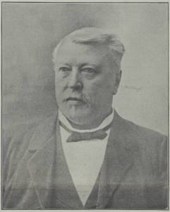 A black-and-white portrait of a middle-aged man. He is shown from the chest up, wearing a formal suit with a patterned vest and bowtie. His hair is neatly combed and he has a mustache., He has a serious, focused expression.