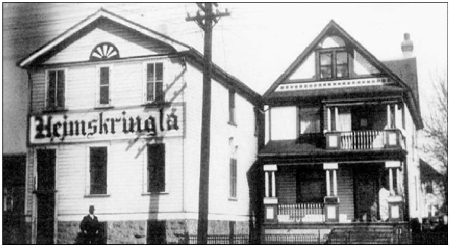 A black and white photo of two early 20th-century two story buildings. The building on the left has five narrows windows and large sign reading Heimskringla. The building on the right looks more like a home with a veranda on the ground floor and a balcony on the second floor.